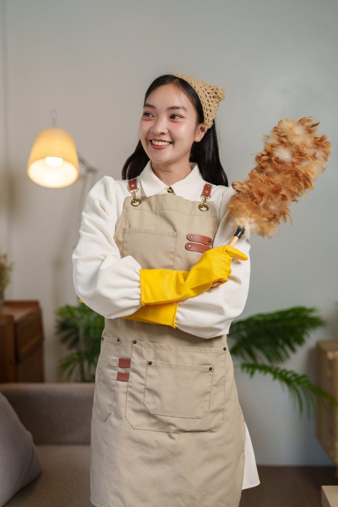 Smiling young housekeeper wearing yellow gloves and an apron holds a feather duster while standing i